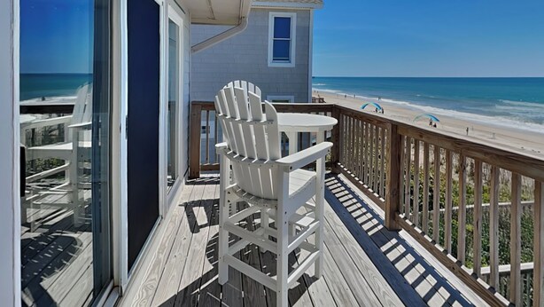 A wooden deck with a white Adirondack chair and a small table, overlooking a beach with a clear blue sky. - Hailey's Hideaway by Topsail Realty (Holly Ridge)