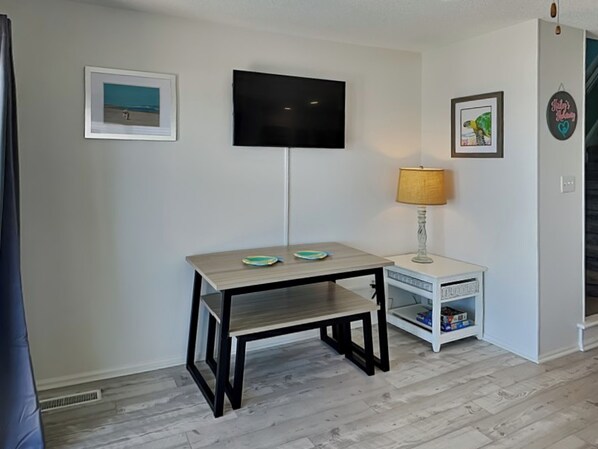 A room with a flat-screen TV mounted on the wall, a small table with a lamp, and a shelf with storage boxes. - Hailey's Hideaway by Topsail Realty (Holly Ridge)