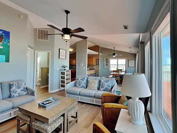 A spacious living room with a sofa, coffee table, and chairs, a dining area with a table and chairs, and a kitchen in the background. - Paradise Palms by Topsail Realty (Holly Ridge)