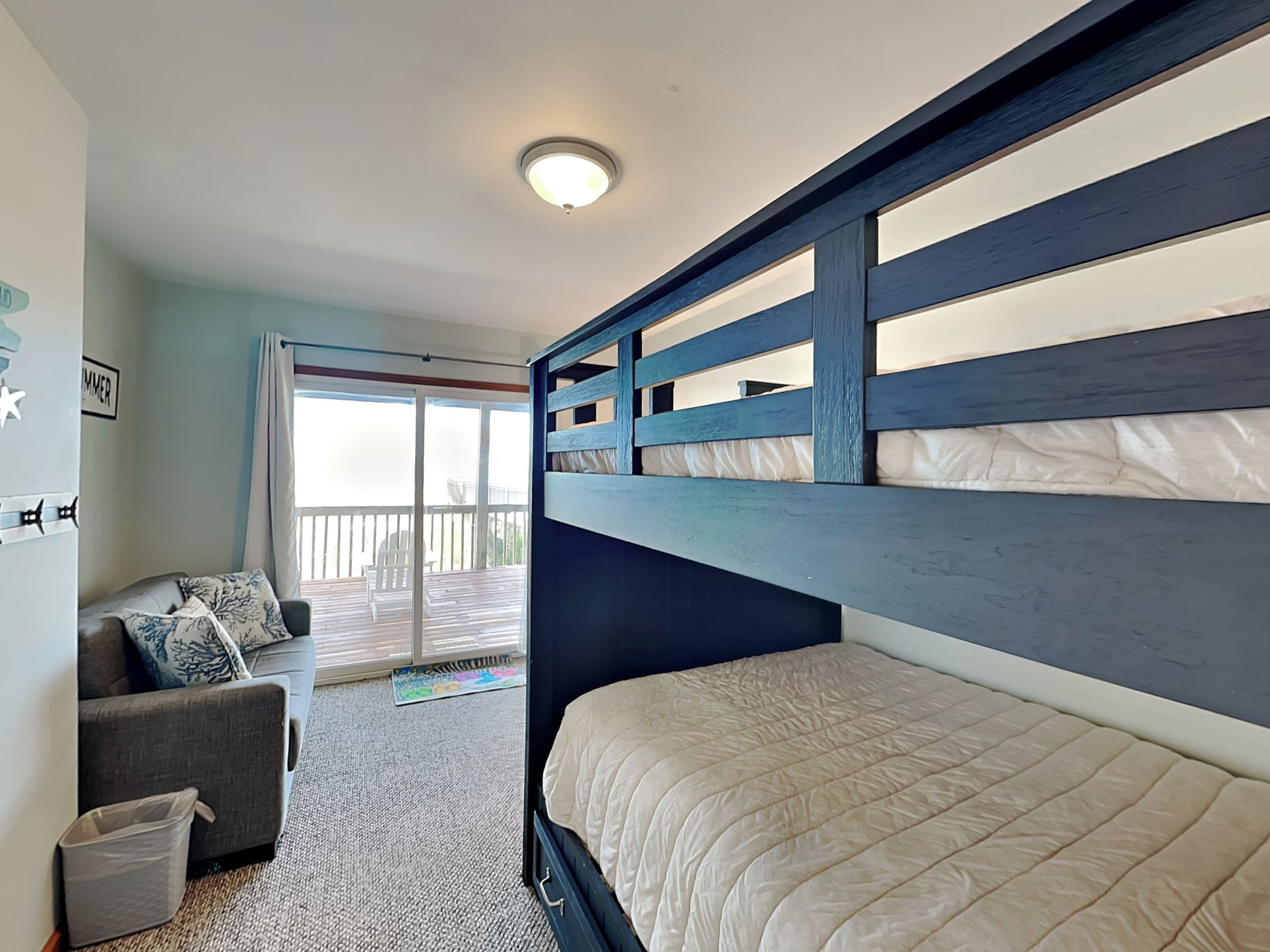 A bedroom with a bunk bed, a sofa, a small trash bin, and a sliding glass door leading to a balcony.