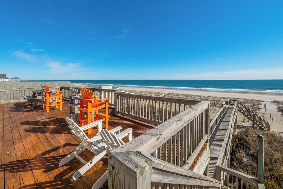 A wooden deck with beach chairs and a wooden railing overlooking a sandy beach and ocean.
