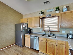A kitchen with wooden cabinets, a stainless steel refrigerator, a dishwasher, and a window with blinds. - Paradise Palms by Topsail Realty (Holly Ridge)