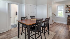 A dining area with a wooden table and chairs, white walls, and a window with blinds. - Jersey Blues by Carolina Retreats (Carolina Beach)