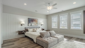 A bedroom with a bed, nightstands, a ceiling fan, and a large window with blinds. - Jersey Blues by Carolina Retreats (Carolina Beach)