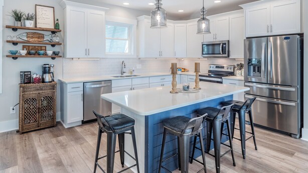A modern kitchen with white cabinets, stainless steel appliances, and a central island with bar stools. - Jersey Blues by Carolina Retreats (Carolina Beach)