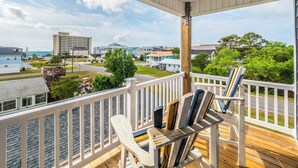 A covered porch with white railings and a ceiling fan, overlooking a residential area with houses and a large building in the distance. - Jersey Blues by Carolina Retreats (Carolina Beach)