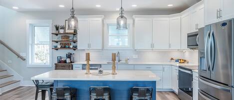 A modern kitchen with white cabinets, a central island with bar stools, stainless steel appliances, pendant lighting, and a staircase leading to another level.