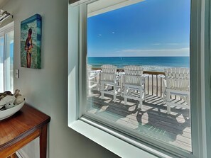 Una habitación con un ventanal grande con vista a una playa, con sillas blancas y una terraza de madera.