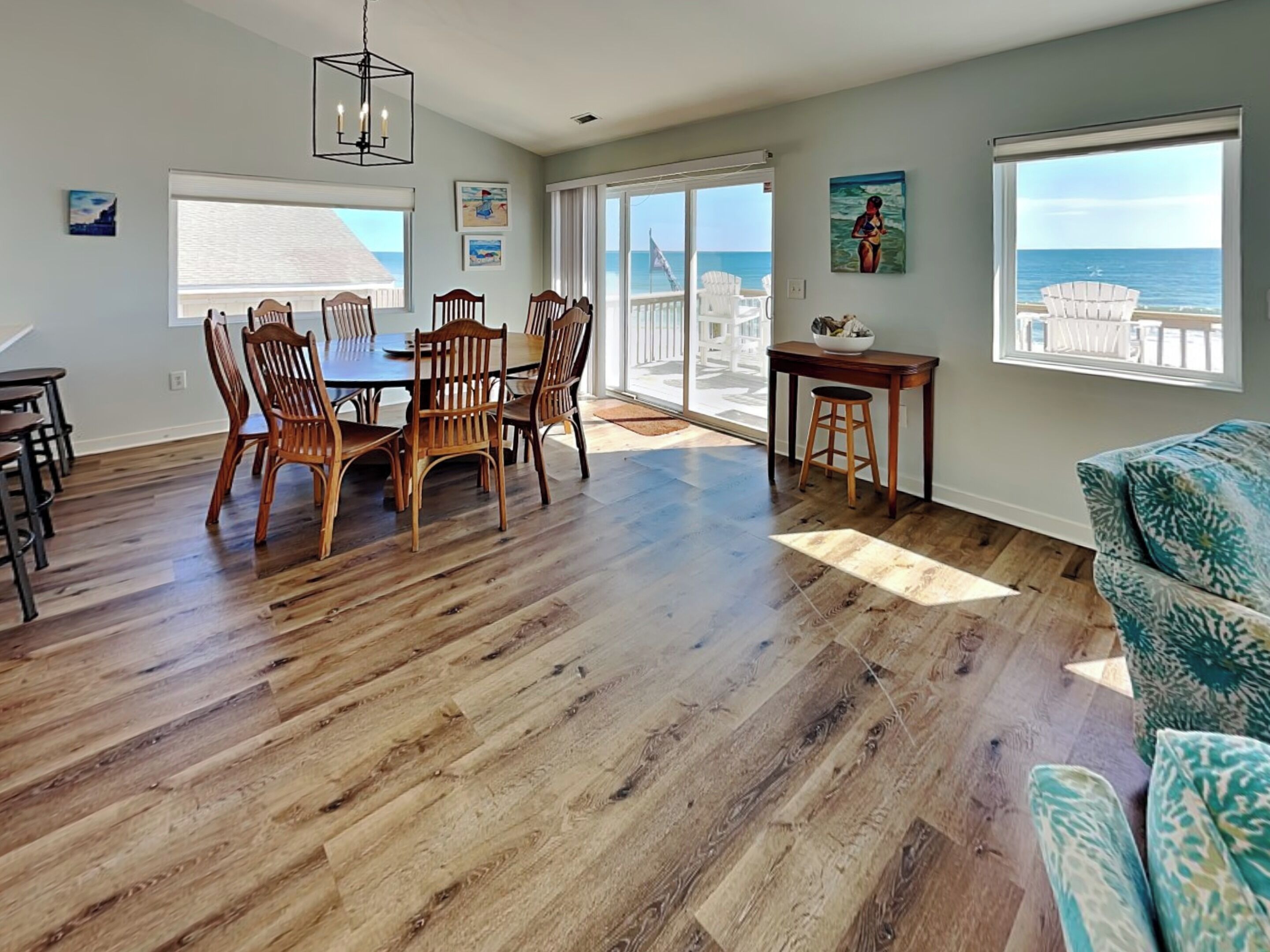 A spacious living room with a wooden floor, a dining table with chairs, a sofa, and a view of the ocean through large windows.