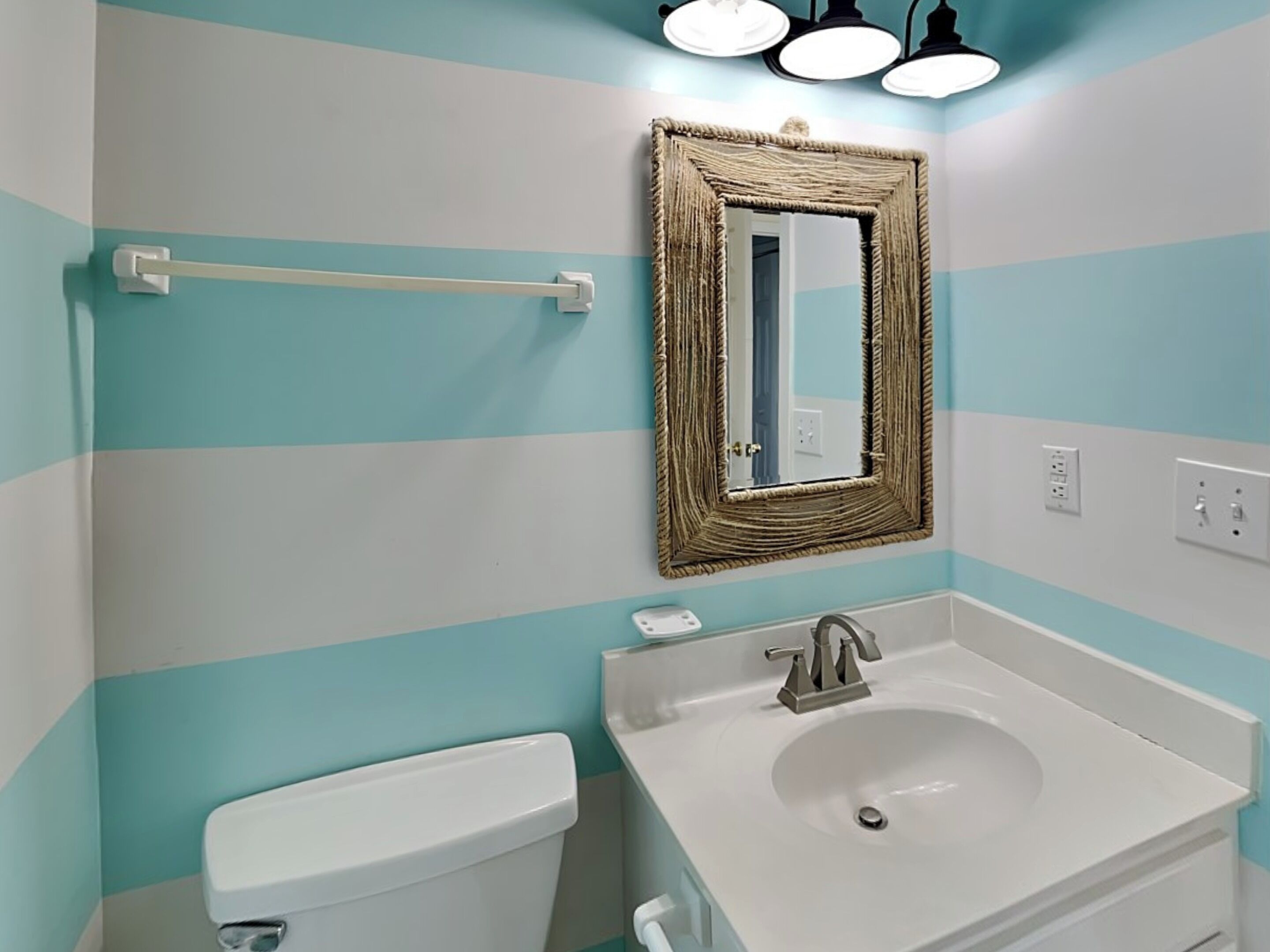 Une salle de bain avec un mur décoré de motifs rayés turquoise et blancs, un lavabo blanc avec un robinet argenté, des toilettes blanches et un miroir avec un cadre tressé.