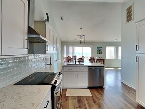 Una cocina moderna con armarios blancos, una estufa negra y una zona de comedor con sillas de madera y vistas al océano.