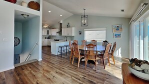 Un comedor amplio con una mesa y sillas de madera, una cocina al fondo y un ventanal que proporciona luz natural.