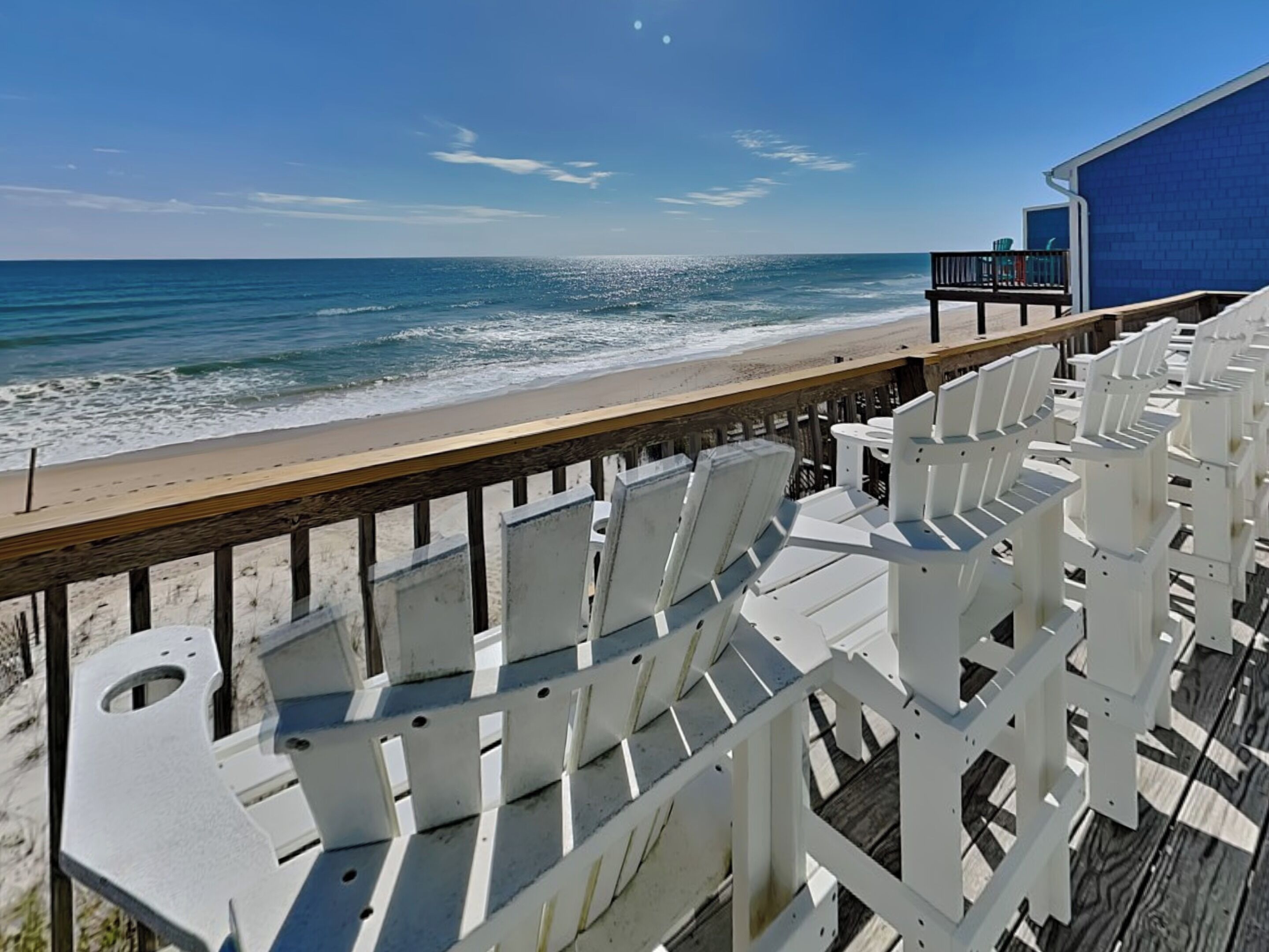 Une terrasse au bord de la plage, avec des chaises blanches et une rambarde en bois donnant sur l’océan.