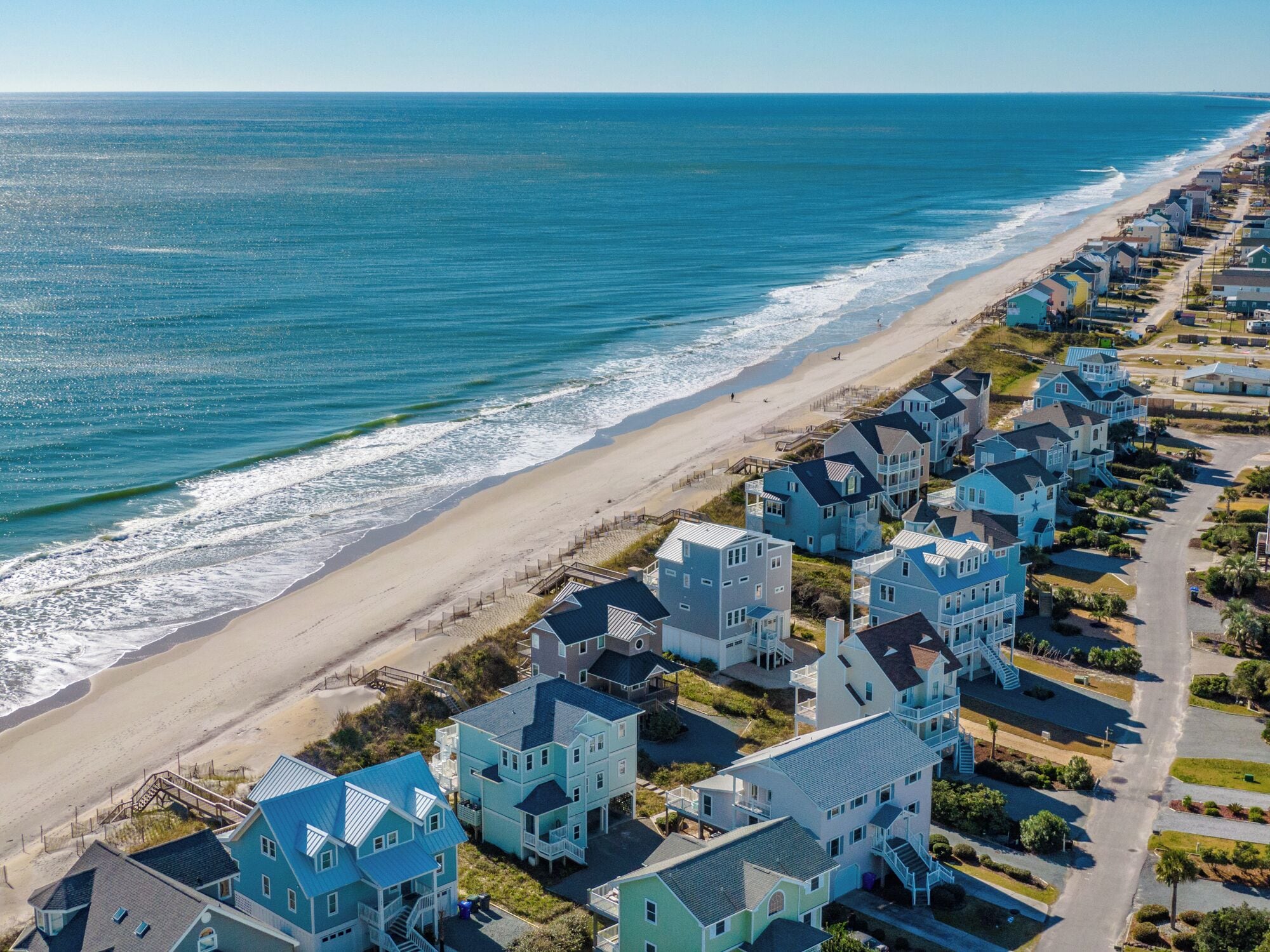 Una zona residencial costera con propiedades frente a la playa, que presenta una mezcla de casas de un solo piso y edificios de varios pisos con balcones, una playa arenosa y el océano al fondo.