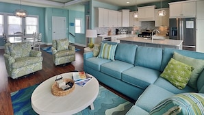 Un salon spacieux avec un canapé bleu, des fauteuils à motifs et une table basse. On aperçoit, au fond, une cuisine avec des meubles blancs et des appareils électroménagers modernes.