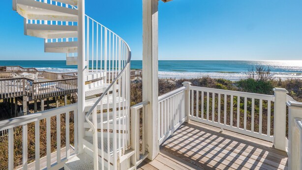Un escalier en colimaçon menant à une terrasse en bois offrant une vue sur la plage, avec des rampes blanches.