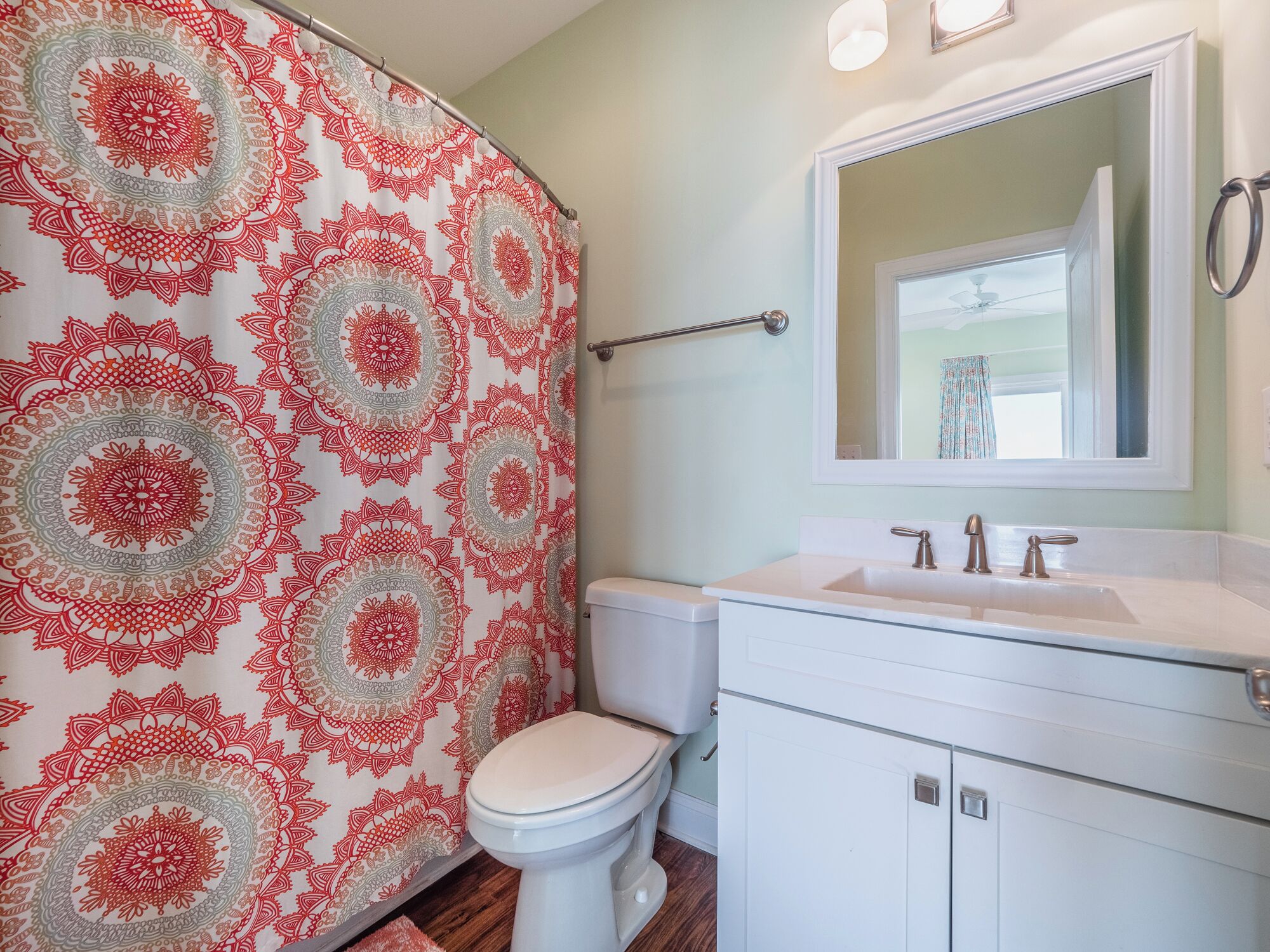 Une salle de bain avec un rideau de douche à motifs, des toilettes blanches, un lavabo avec un miroir au-dessus et un plancher en bois.