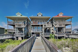 A beachfront property with multiple balconies, a wooden deck leading to the entrance, and a clear sky.