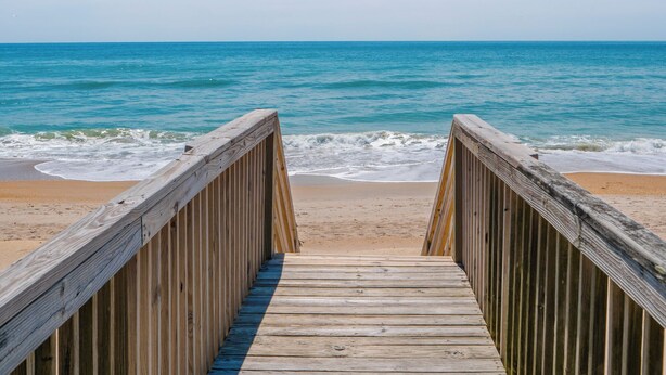 Une promenade en bois qui mène à une plage où l’eau est d’un bleu limpide et le rivage est sablonneux.