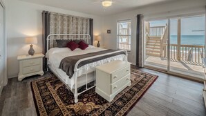 Une chambre à coucher comprenant un lit, des tables de chevet, une lampe de chevet, un ventilateur de plafond, une fenêtre avec des stores et une vue sur la plage depuis un balcon.