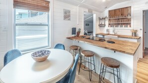 Une cuisine moderne avec des meubles blancs, un comptoir en bois et des tabourets de bar. On y trouve une fenêtre avec des stores, un bol sur la table et une enseigne indiquant « Seaside Bakery ».
