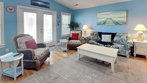 A living room with a blue wall, two windows with blinds, a white coffee table, two patterned sofas, and a small side table with a lamp. - No Worries in Paradise (Carolina Beach)