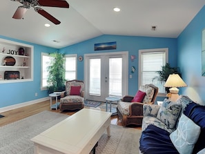 A living room with a ceiling fan, a white coffee table, two wicker chairs, a blue sofa, and a bookshelf with decorative items. - No Worries in Paradise (Carolina Beach)