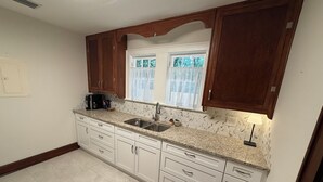 Una cocina con armarios blancos, encimera de granito, pileta doble y ventana con cortinas traslúcidas.