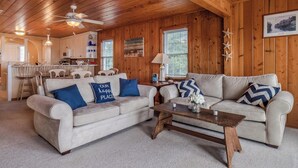 A cozy living room with two couches, a coffee table, wooden walls, and a kitchen in the background.