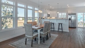 A modern kitchen with white cabinets, a stainless steel refrigerator, and a dining area with wicker chairs and a wooden table. - Happily Ever After by Topsail Realty (Holly Ridge)