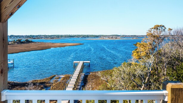 A wooden deck with a view of a calm body of water and a distant shoreline. - Happily Ever After by Topsail Realty (Holly Ridge)