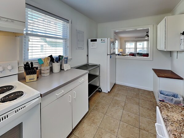 A kitchen with white cabinets, a stove, a sink, and a window with blinds. - Good Vibrations by Carolina Retreats (Carolina Beach)