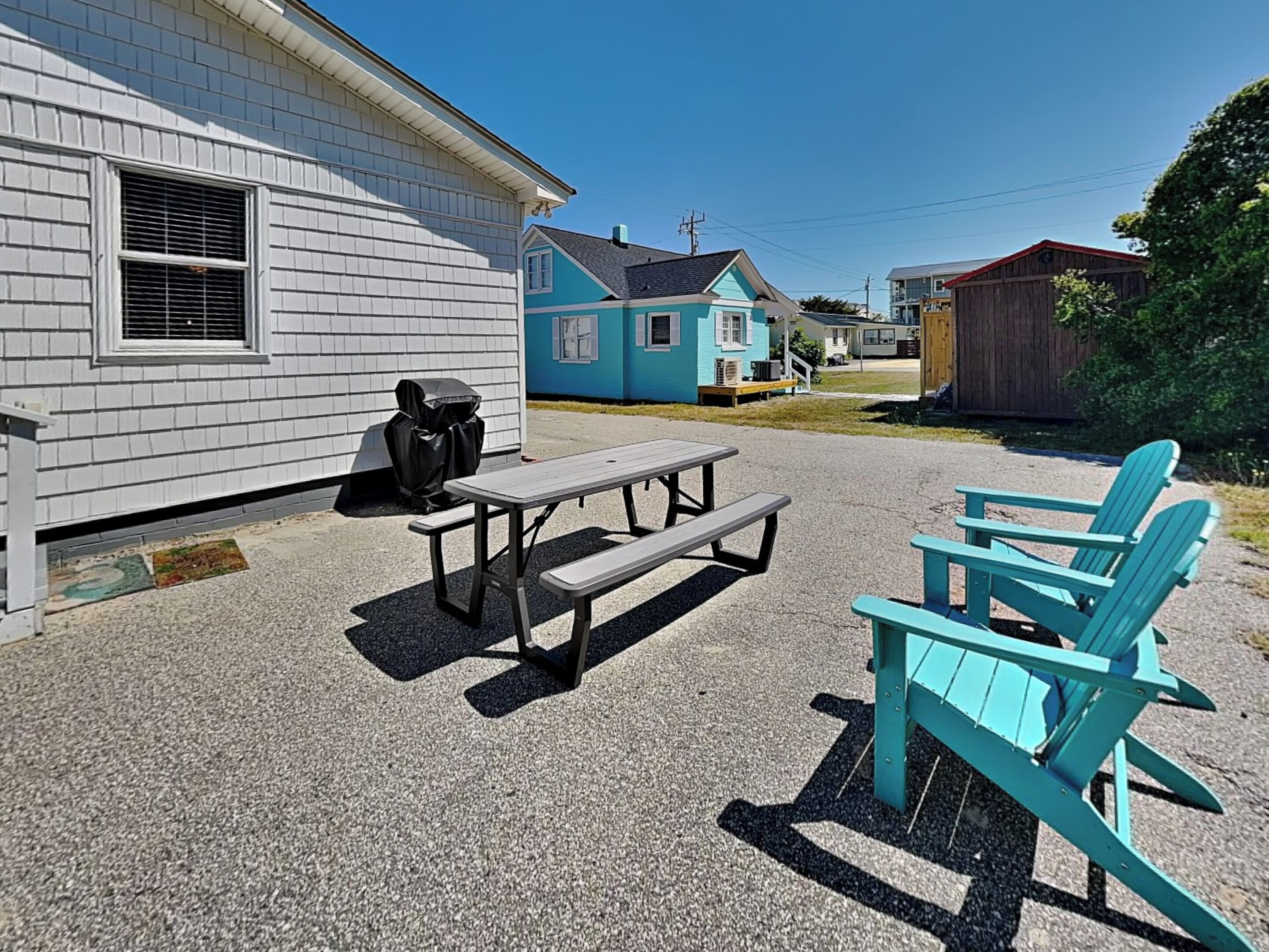 A picnic area with a table and chairs, a covered grill, and a house in the background.