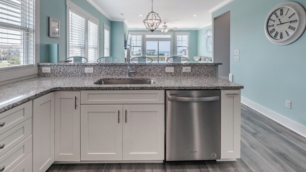 A modern kitchen with a granite countertop, stainless steel dishwasher, and light blue walls. - Strand Bungalow by Topsail Realty (Holly Ridge)