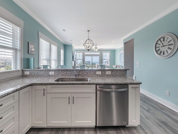 A modern kitchen with a granite countertop, stainless steel dishwasher, and light blue walls. - Strand Bungalow by Topsail Realty (Holly Ridge)