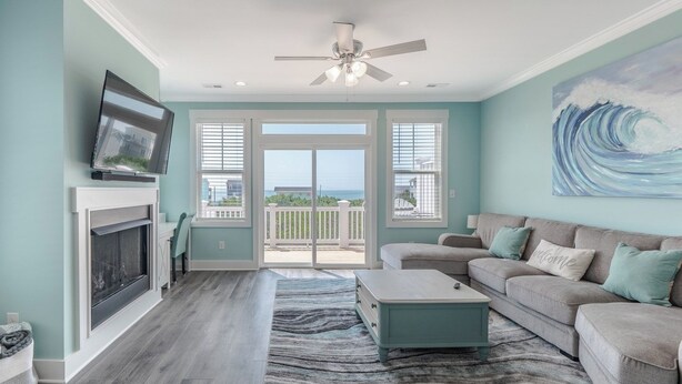 A spacious living room with a fireplace, a television, a ceiling fan, and a large window offering a view of the ocean.