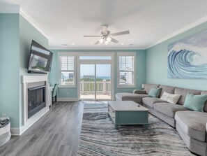 A spacious living room with a fireplace, a television, a ceiling fan, and a large window offering a view of the ocean. - Strand Bungalow by Topsail Realty (Holly Ridge)