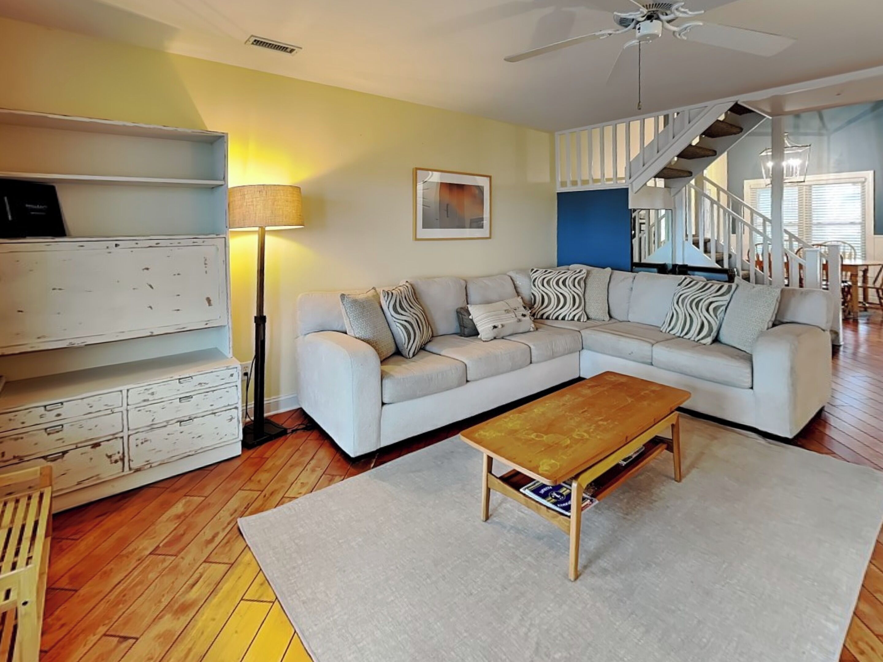A living room with a sectional sofa, a coffee table, a lamp, a bookshelf, and a staircase.