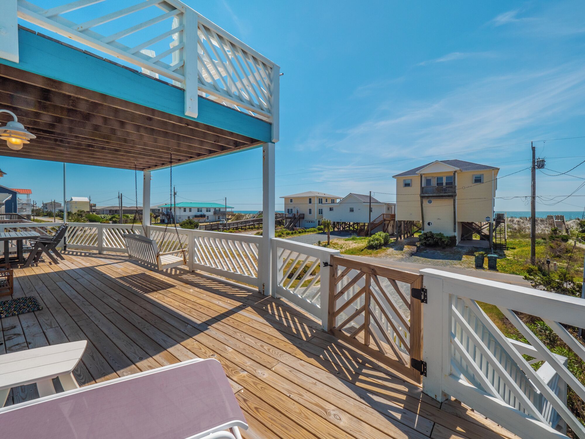 Une terrasse en bois avec des meubles d’extérieur, un espace couvert doté d’une rambarde blanche, et une vue sur les maisons et la plage en arrière-plan.