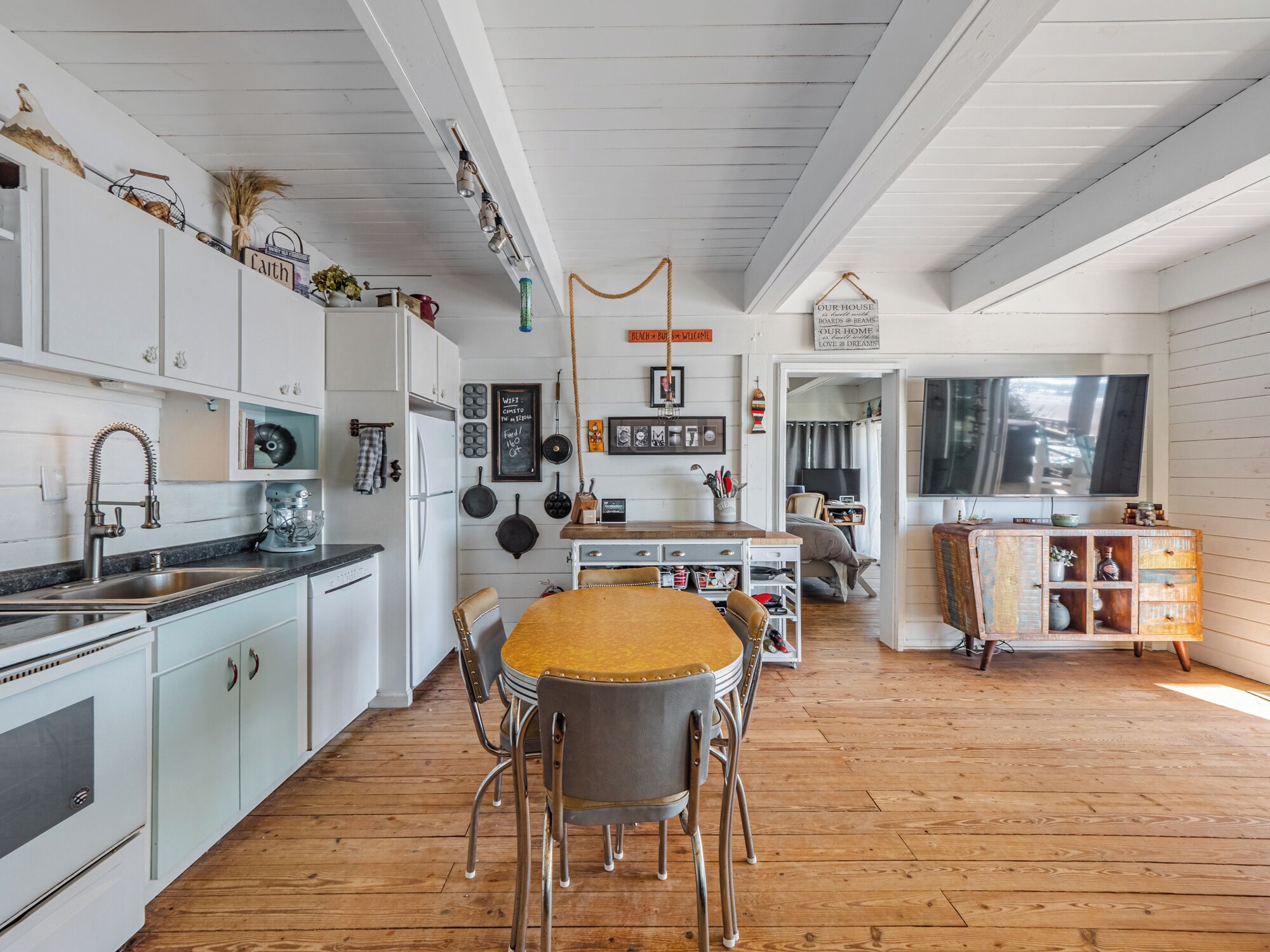 A kitchen with white cabinets, a wooden dining table with four chairs, a hanging light fixture, and a large window.