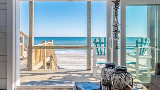 A beach view from a room with a large window, wooden deck, and two beach chairs. - Vintage Vacation by Topsail Realty (Holly Ridge)