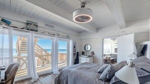 A bedroom with a large bed, a desk, and a view of the beach through a sliding glass door. - Vintage Vacation by Topsail Realty (Holly Ridge)