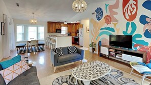 A modern living room with a sofa, coffee table, and TV. There is a kitchen in the background with wooden cabinets and a dining area with chairs. - Sea Angels by Topsail Realty (Holly Ridge)