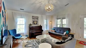 A modern living room with a piano, a sofa, a television, and a decorative rug. - Sea Angels by Topsail Realty (Holly Ridge)