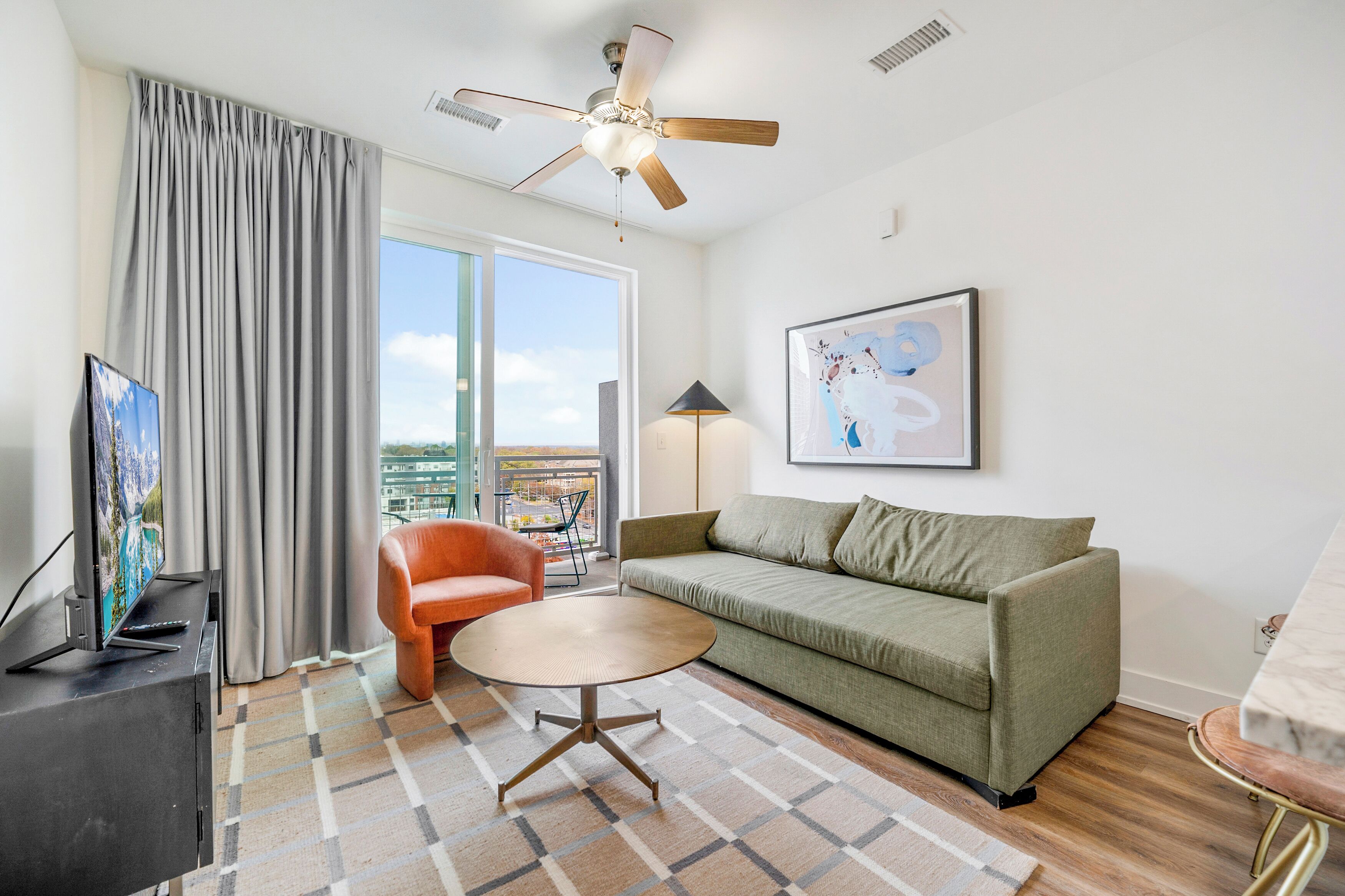 Un salon moderne avec un canapé vert, une table basse ronde, un ventilateur de plafond, un téléviseur et une grande fenêtre avec des rideaux.