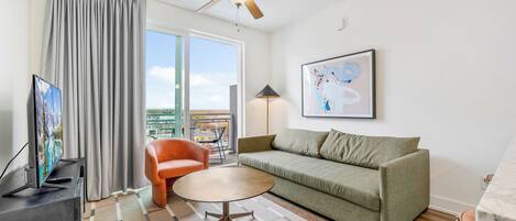 Un salon moderne avec un canapé vert, une table basse ronde, un ventilateur de plafond, un téléviseur et une grande fenêtre avec des rideaux.