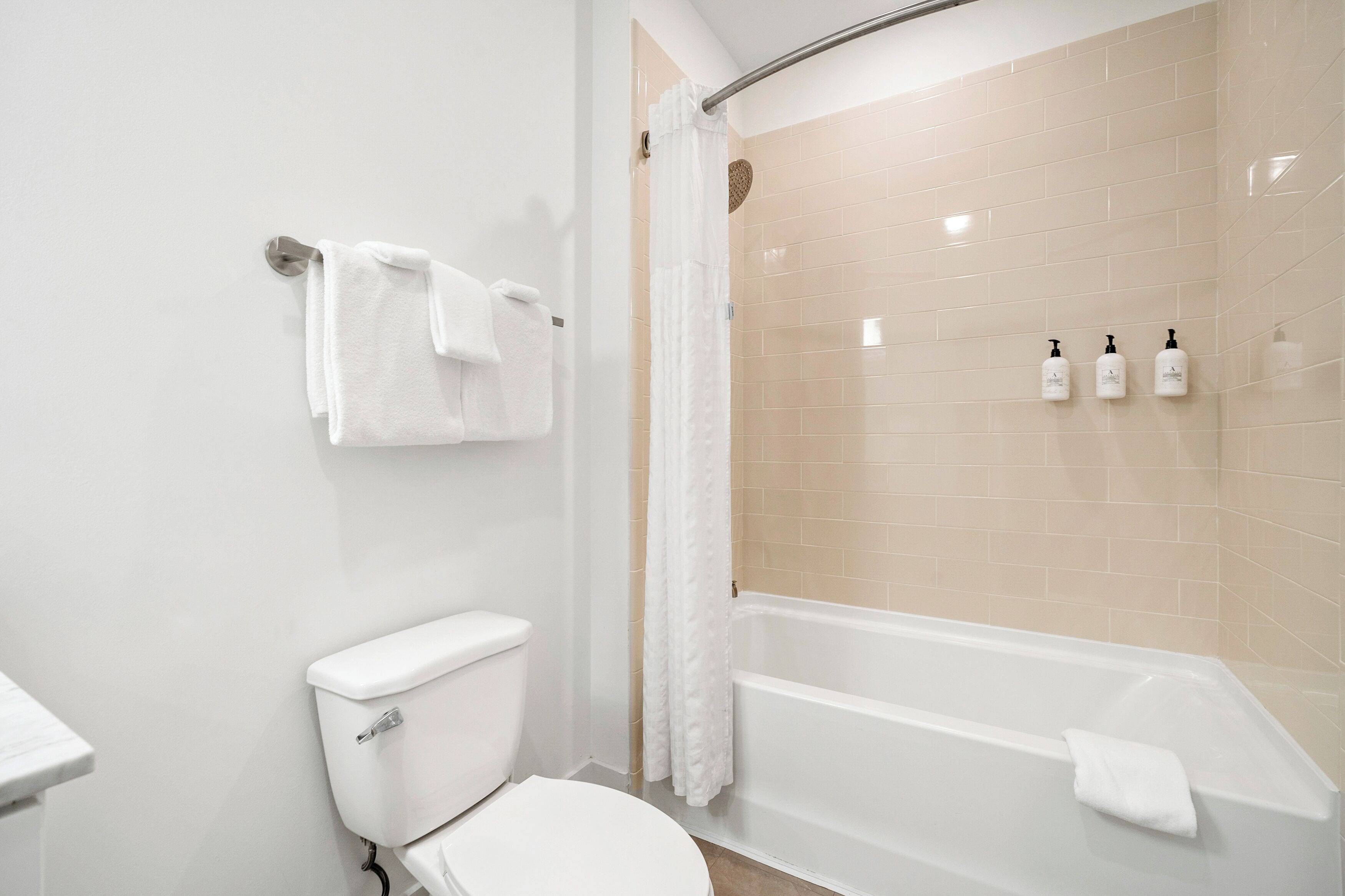 A bathroom with a toilet, a bathtub, and a shower with beige tiles. There are three bottles on the wall.