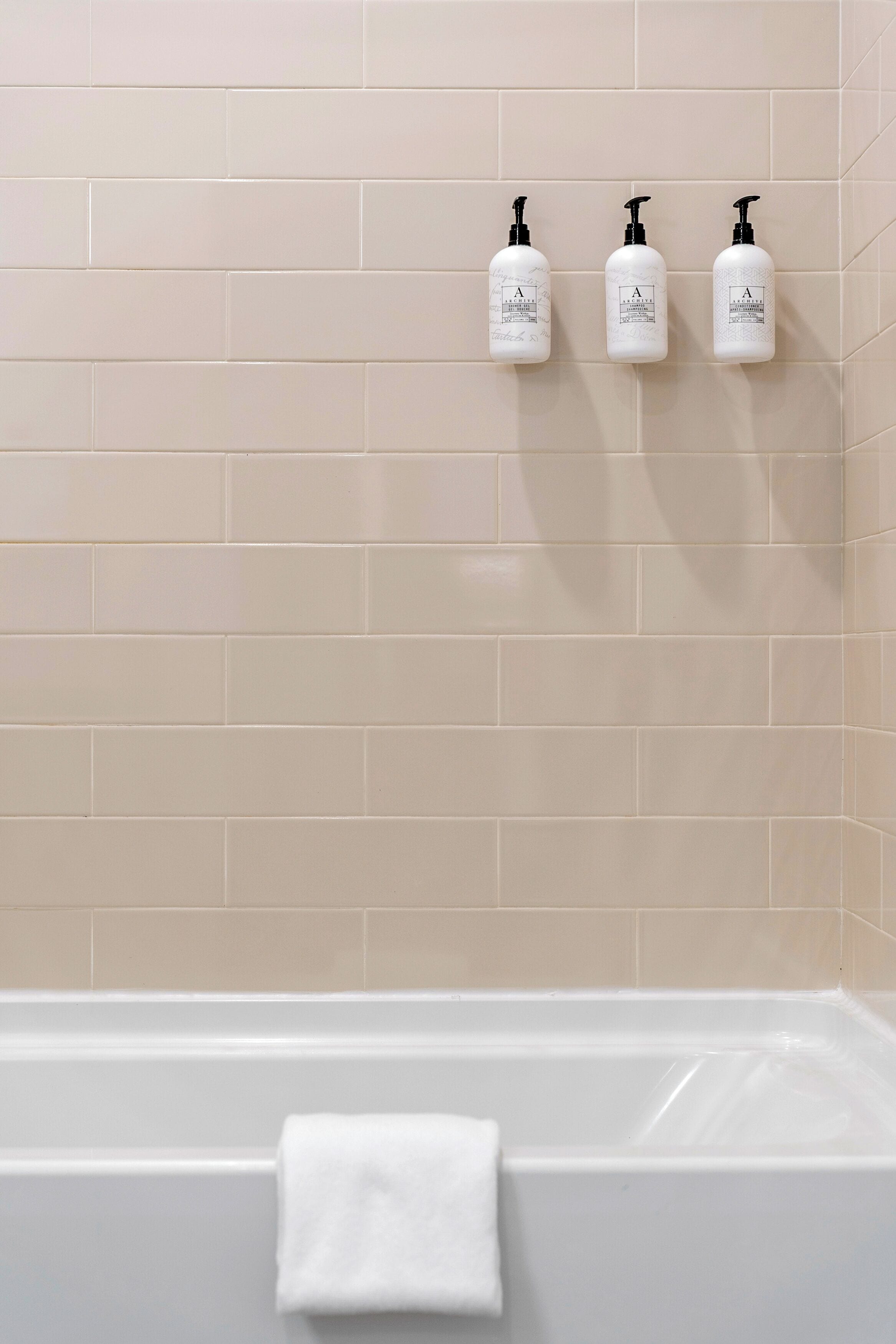 Un baño con paredes de baldosas color beige, una bañera blanca y tres botellas blancas con dispensadores negros en la pared.