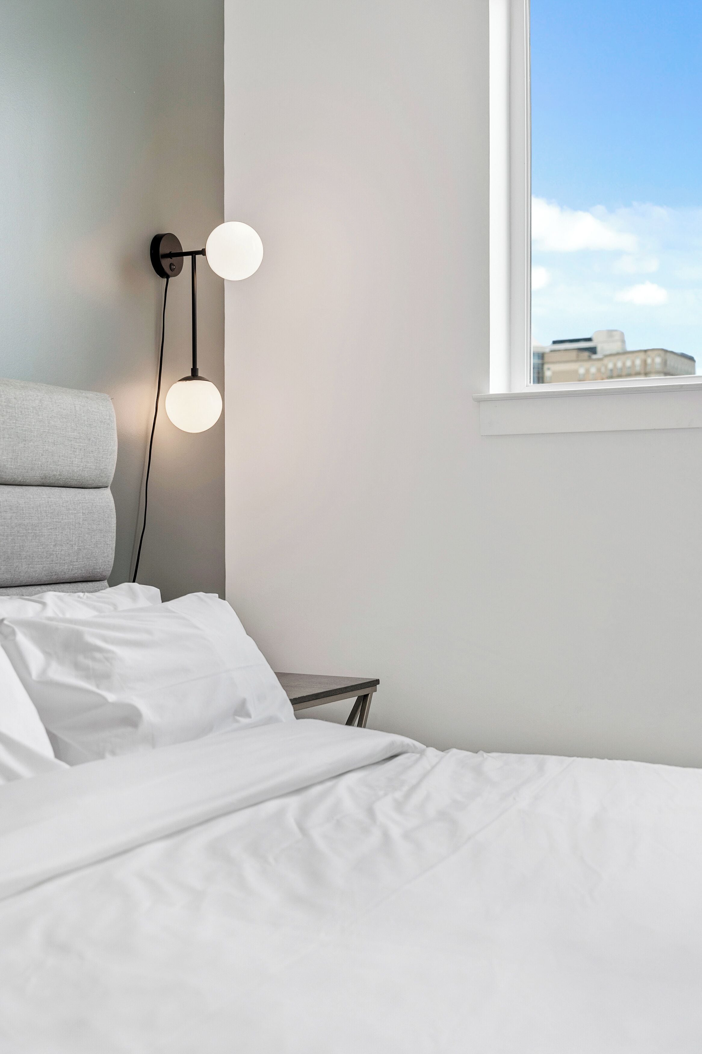 A bedroom with a bed, a nightstand, a wall-mounted lamp, and a window with a view of buildings.