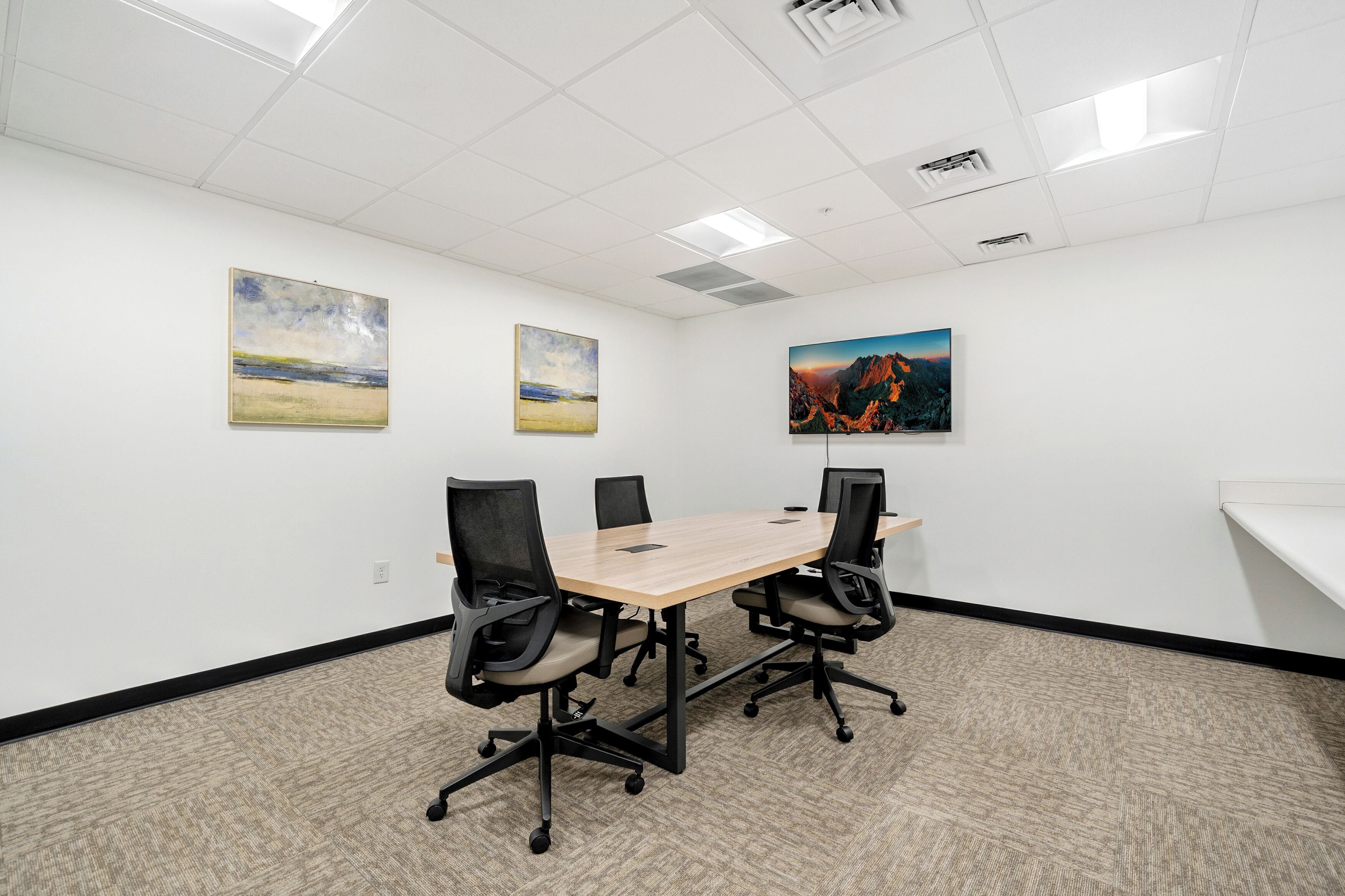 A conference room with a wooden table, four black office chairs, and three paintings on the walls.